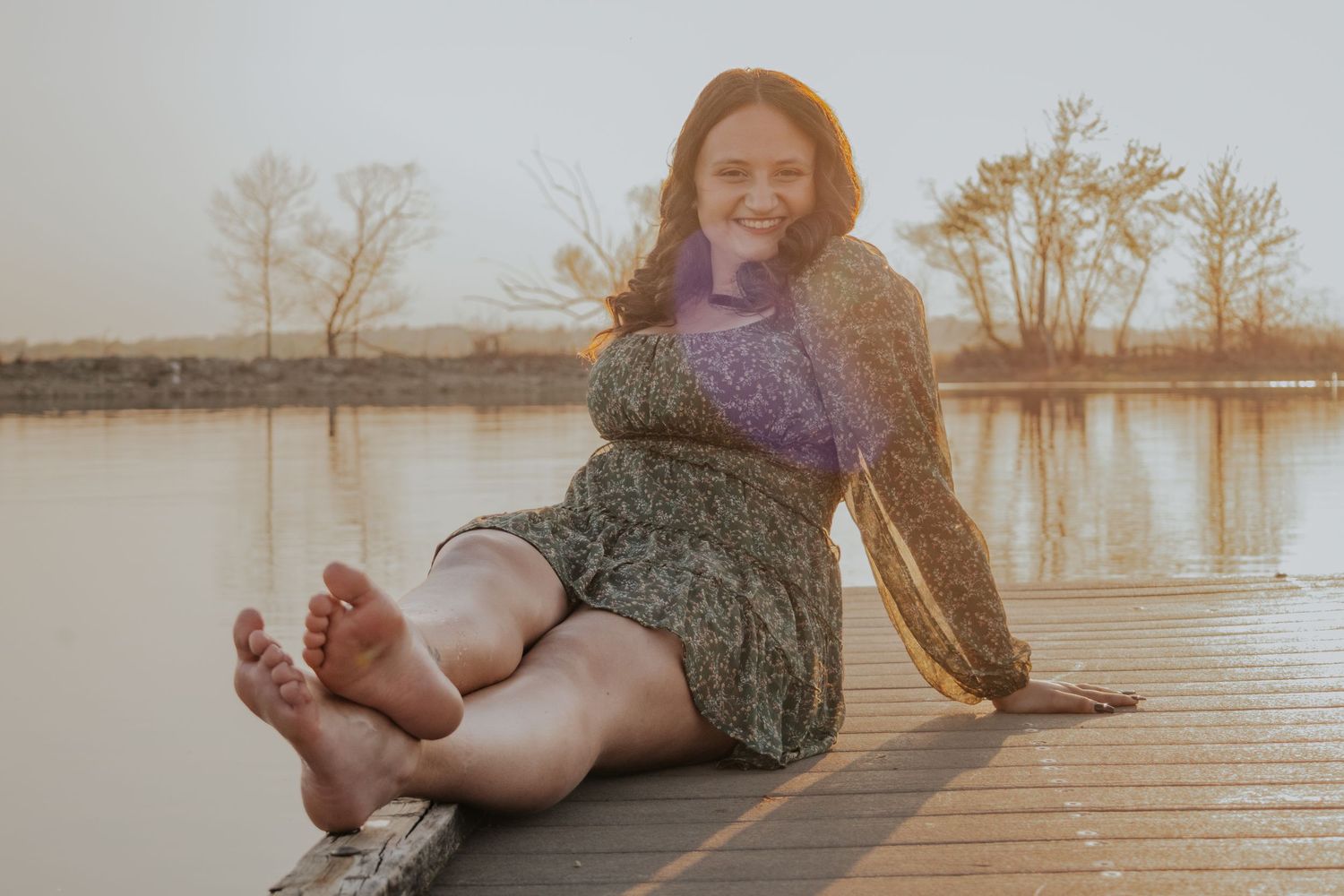 Someone wearing a purple sweater and patterned dress sits barefoot on a wooden dock by a tranquil lake at sunset.