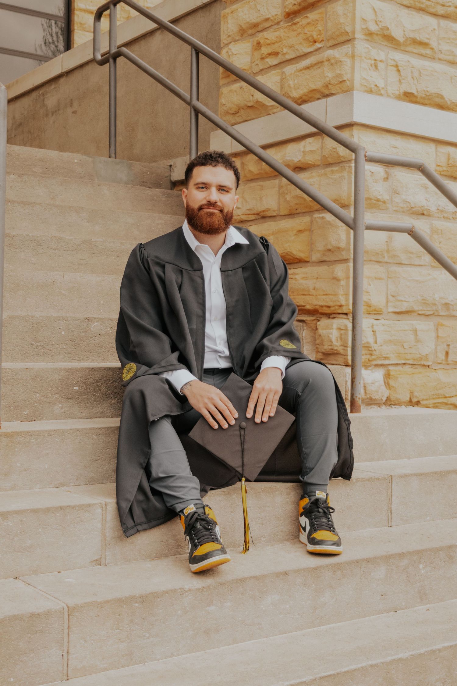 Someone with a ginger beard sits casually on stone steps wearing a black jacket, gray pants and yellow-accented sneakers.