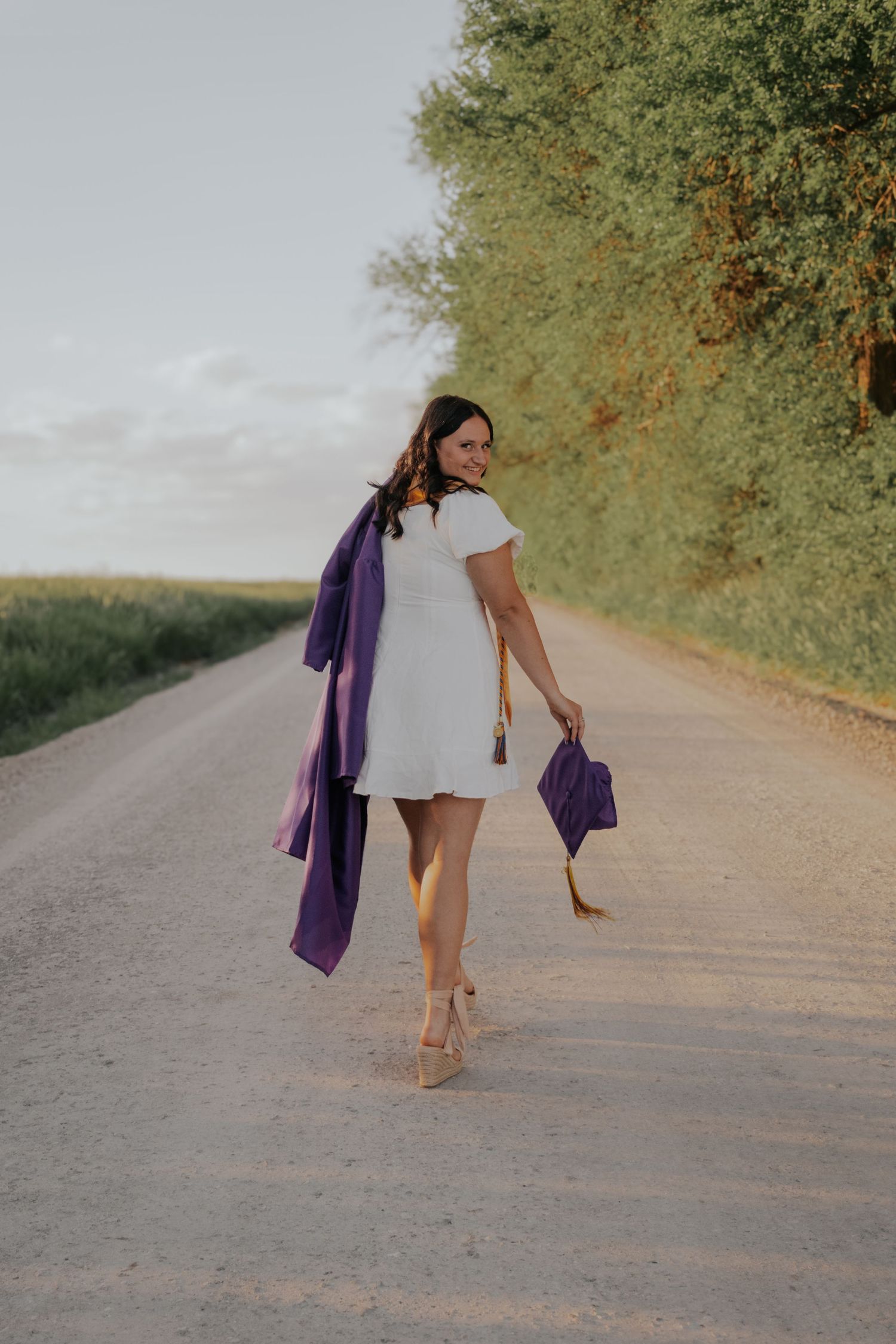 Woman in white dress walking on rural road with purple accessories and heels at golden hour.