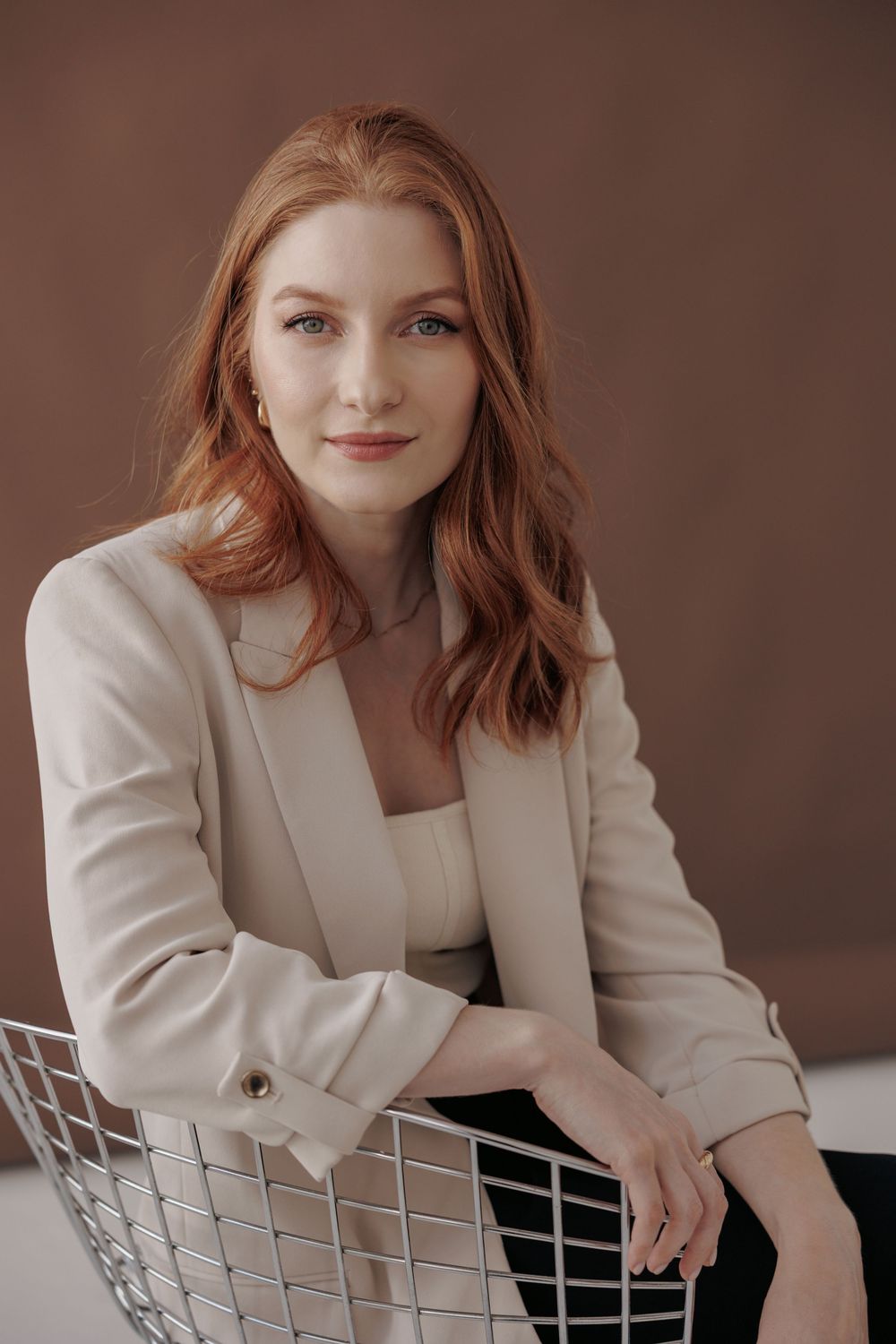 Professional woman in cream blazer leans thoughtfully on white decorative grid in studio setting.