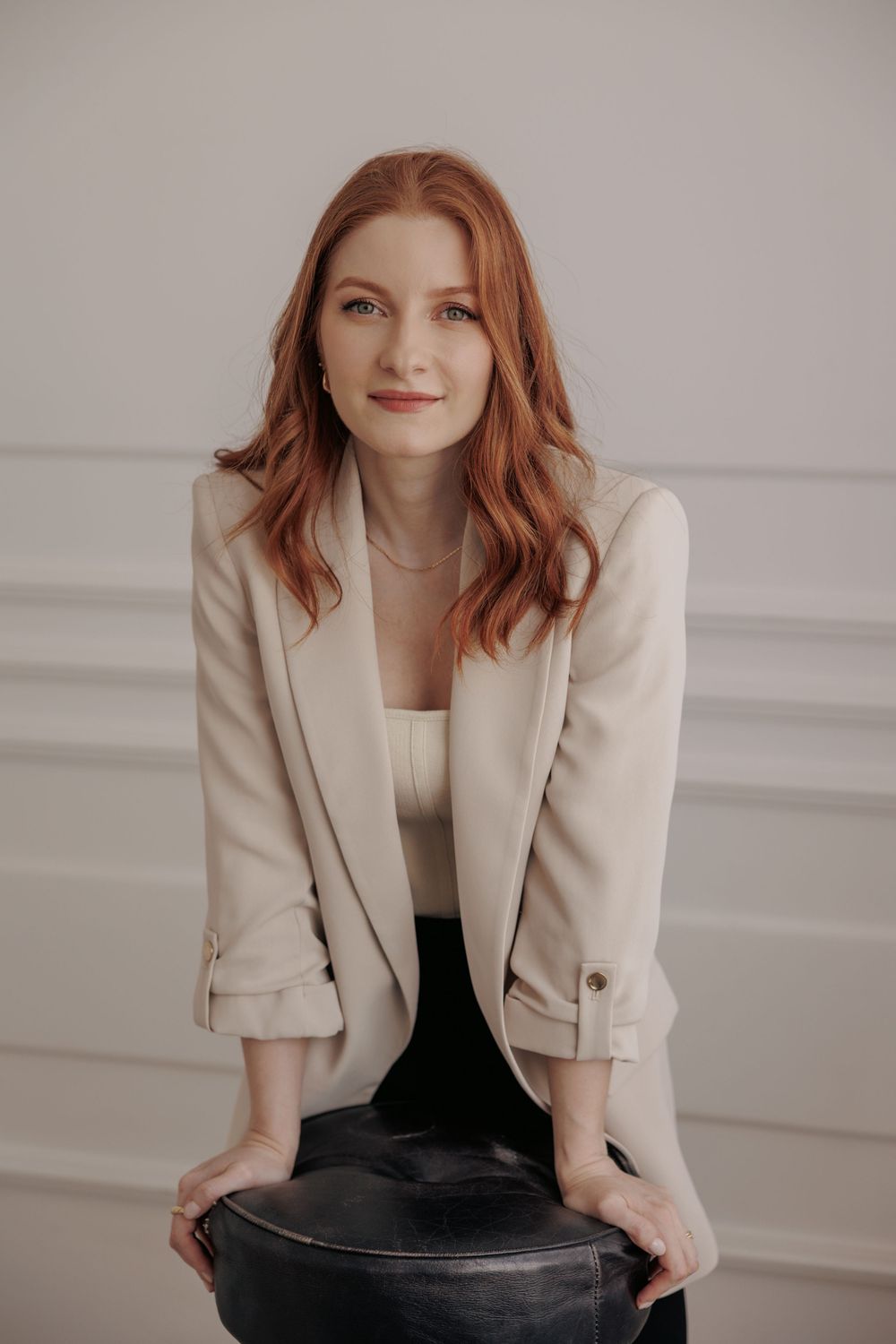 Professional portrait of someone with wavy red hair wearing a beige blazer and sitting on a dark chair against a light wall.