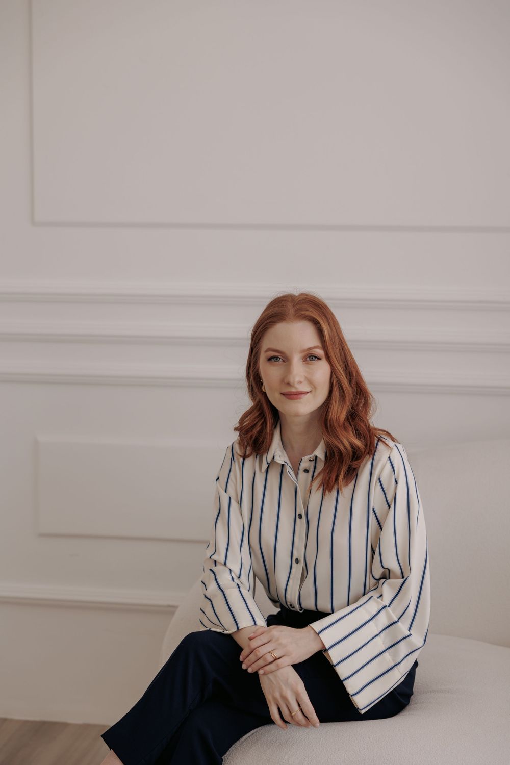 A person with red hair sits on a white couch wearing a striped white button-down blouse and dark pants.