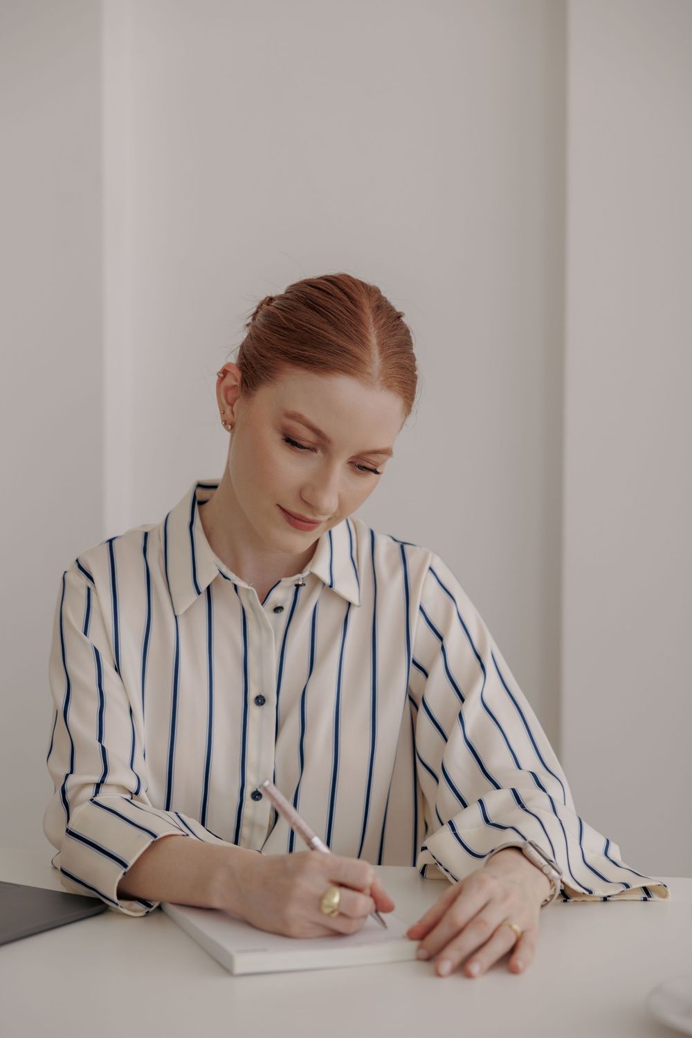 A person in a striped white and blue button-down shirt writes in a notebook while seated at a white desk.
