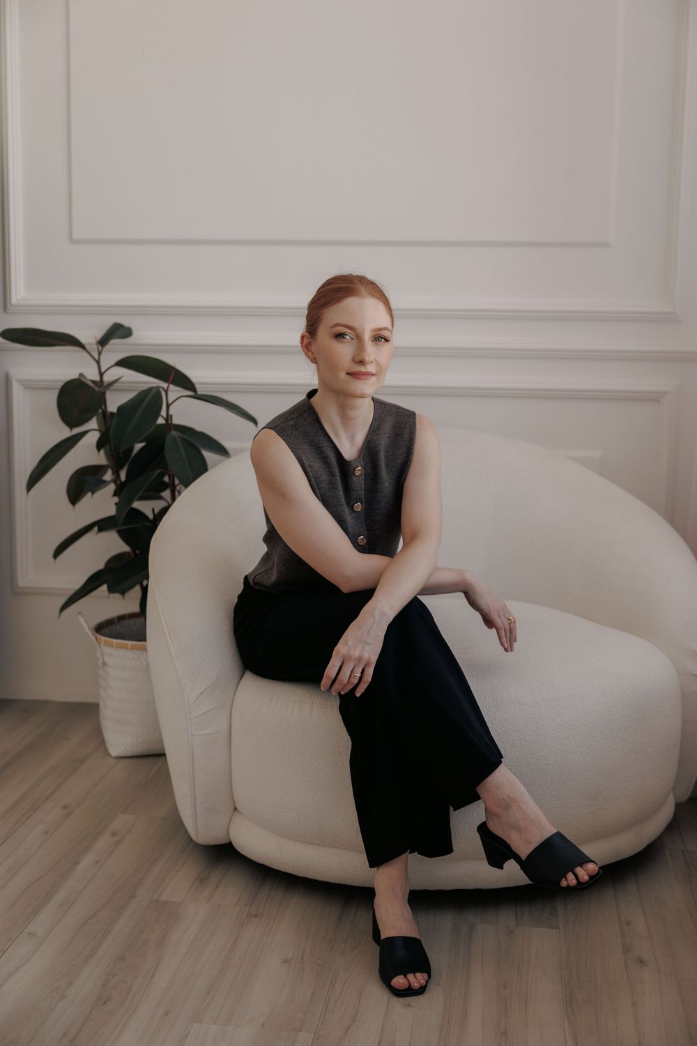 A figure in a black dress sits elegantly on a white modern chair against a light wall with indoor plants in view.