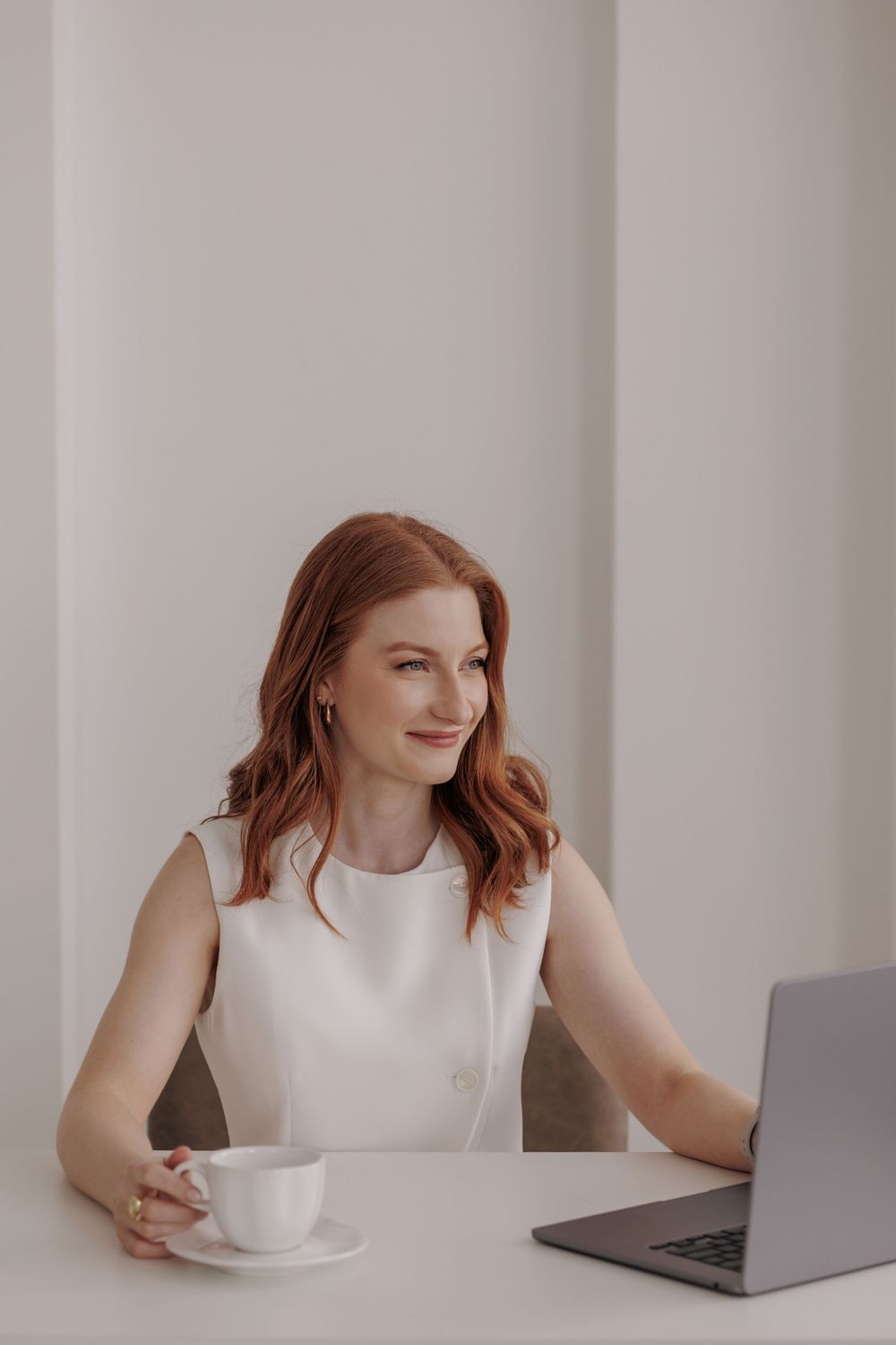 Professional in white sleeveless top working at laptop while seated at minimalist white desk with coffee cup.