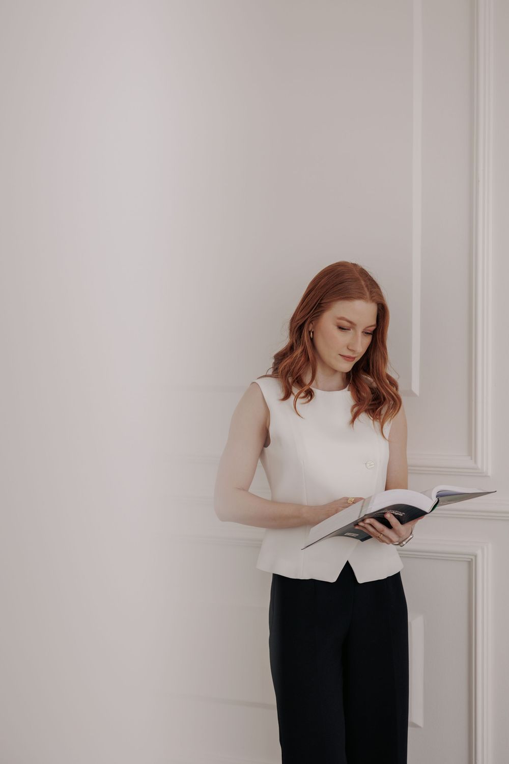 Person in black attire reads from a white book against a light wall.