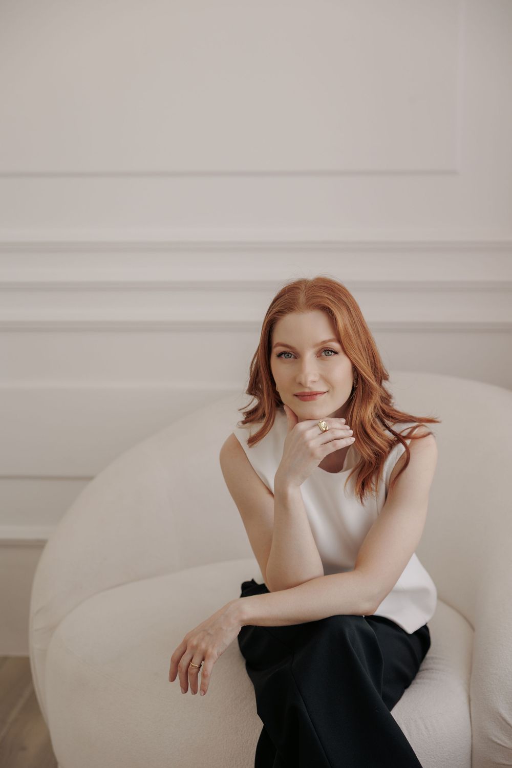A person with red hair sits casually on a white chair wearing a white top and black pants in a minimalist setting.