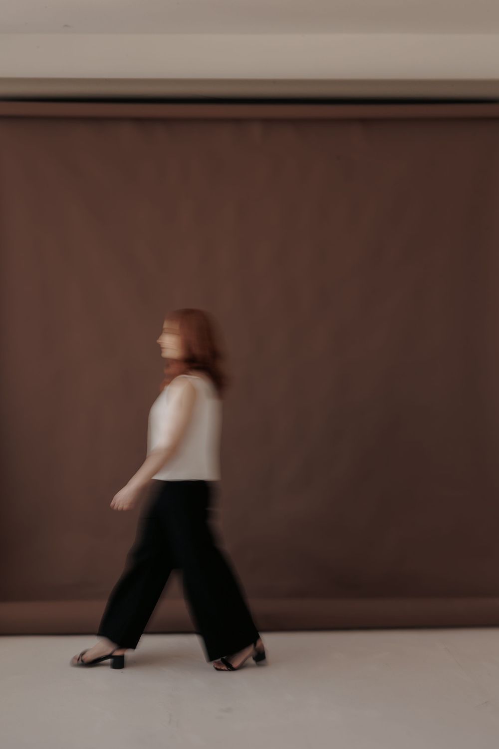 Motion-blurred figure in white top and black wide-leg pants walking against a brown studio backdrop.