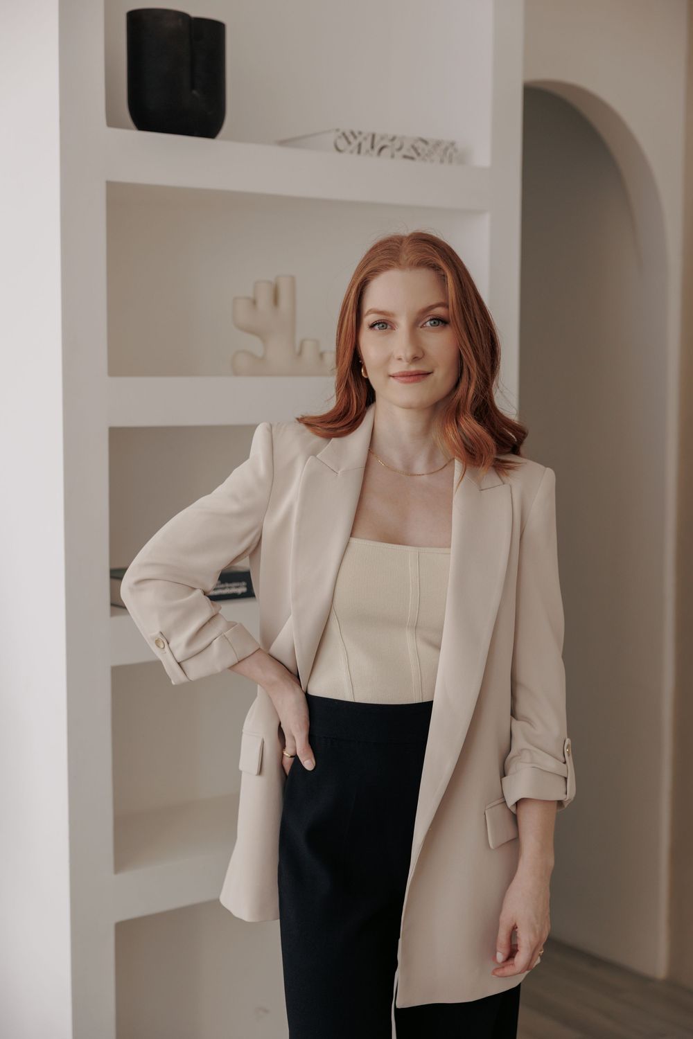 Professional studio fashion photography of a woman wearing a light beige blazer and black top in white interior setting.