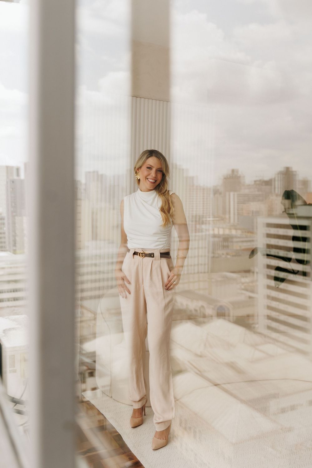 Person in monochromatic cream outfit poses in modern high-rise building with city skyline view through columns.