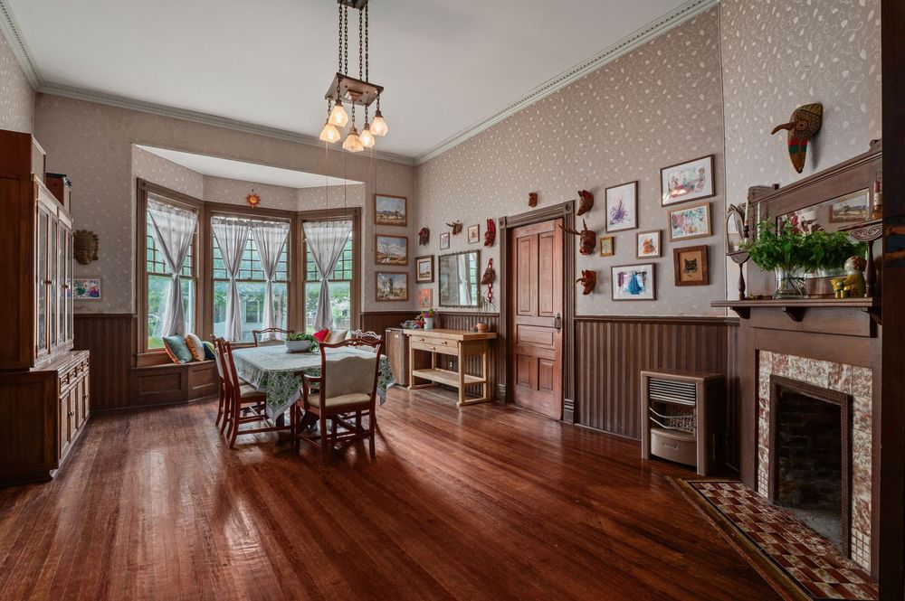 Victorian era dining room with bay windows, hardwood floors, ornate fireplace and vintage framed artwork on wallpapered walls.