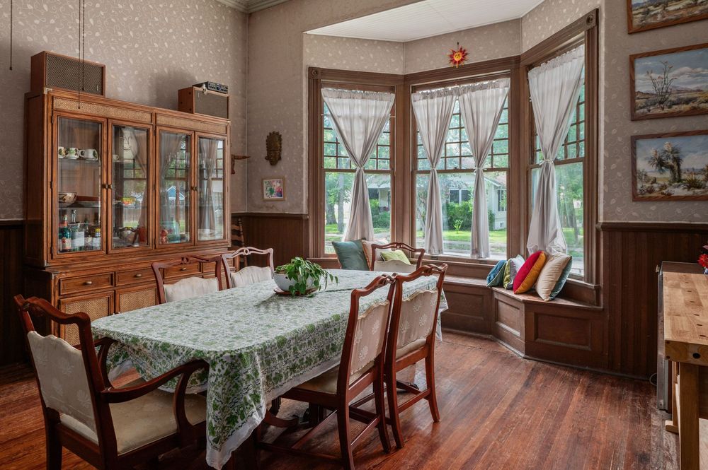 Dining room with wooden wainscoting, patterned wallpaper, and large windows letting in natural light.