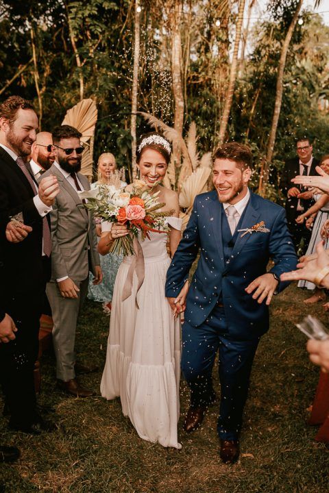 Newlyweds walk through a wooded outdoor ceremony as guests throw confetti in celebration.