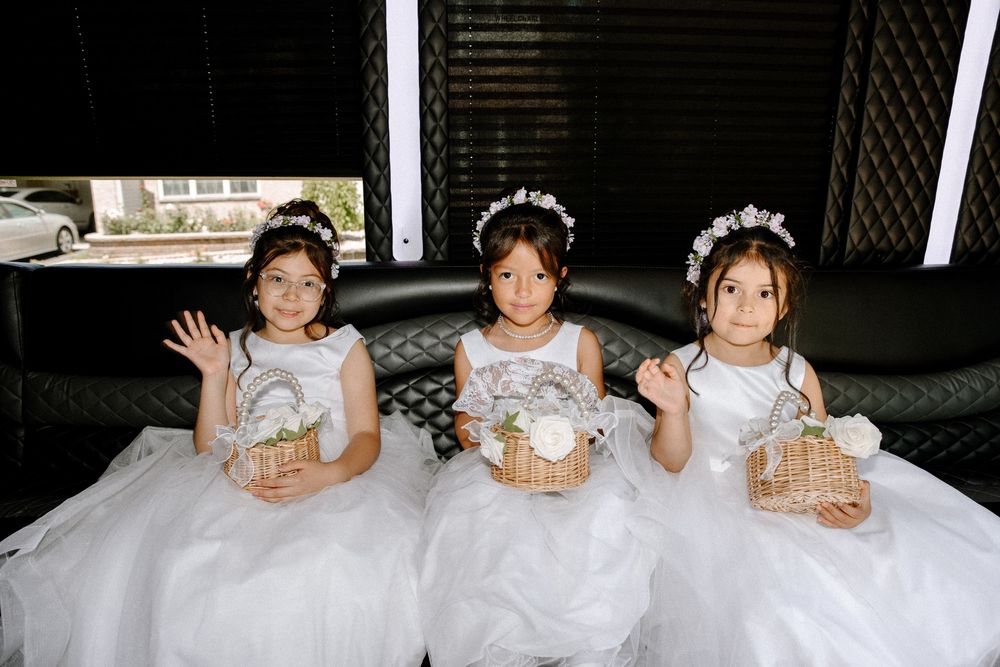 Three young flower girls in white dresses sit together holding baskets and waving at the camera.