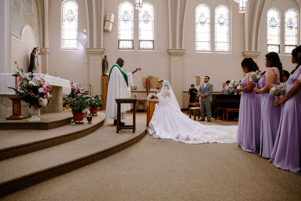 Church wedding ceremony with bridesmaids in purple dresses and white floral arrangements at the altar.
