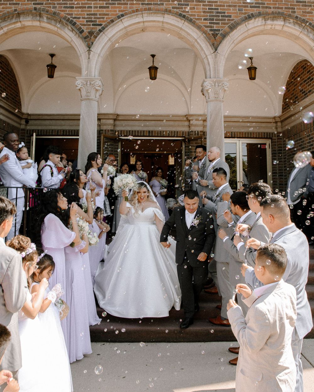 Wedding party celebrates under ornate brick archways with bubbles and confetti.