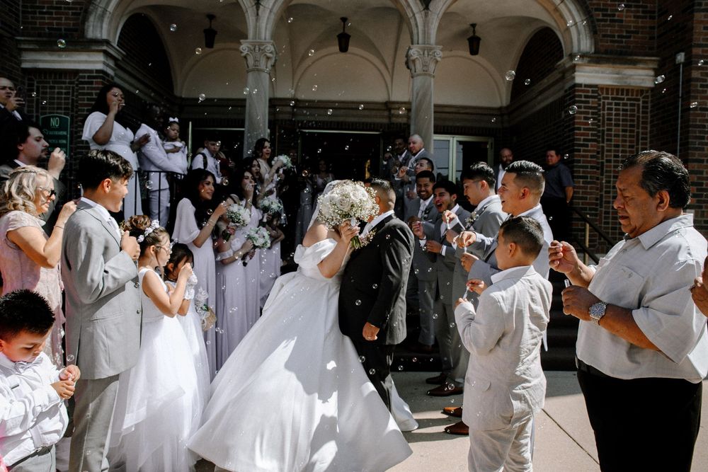 Wedding guests and wedding party dressed in white gather for a special moment outdoors at a brick venue.