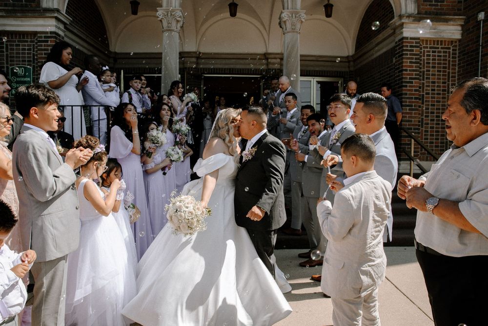 Wedding party gathered outside brick building in white and gray attire.