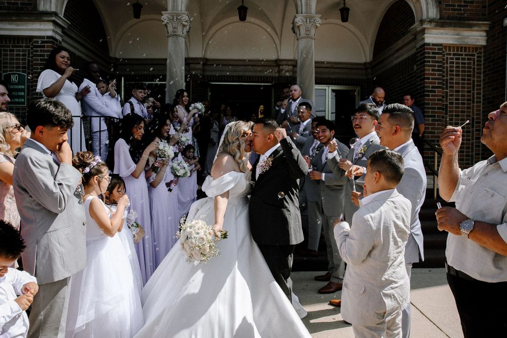 Large wedding party in crisp white dresses and suits pose on brick-columned steps of venue.