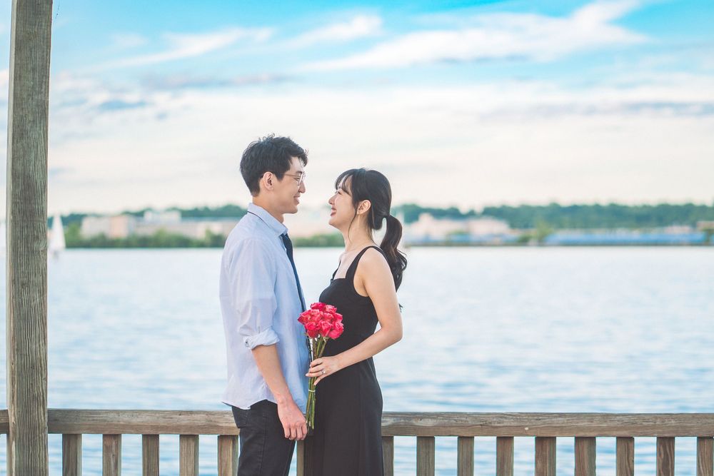 A romantic couple stands together on a waterfront deck holding red flowers against a serene blue water backdrop.