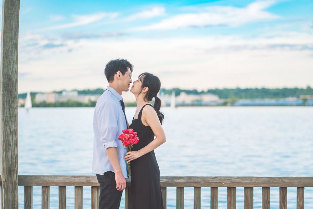A couple embraces on a waterfront deck with red flowers and a scenic harbor view during sunset.