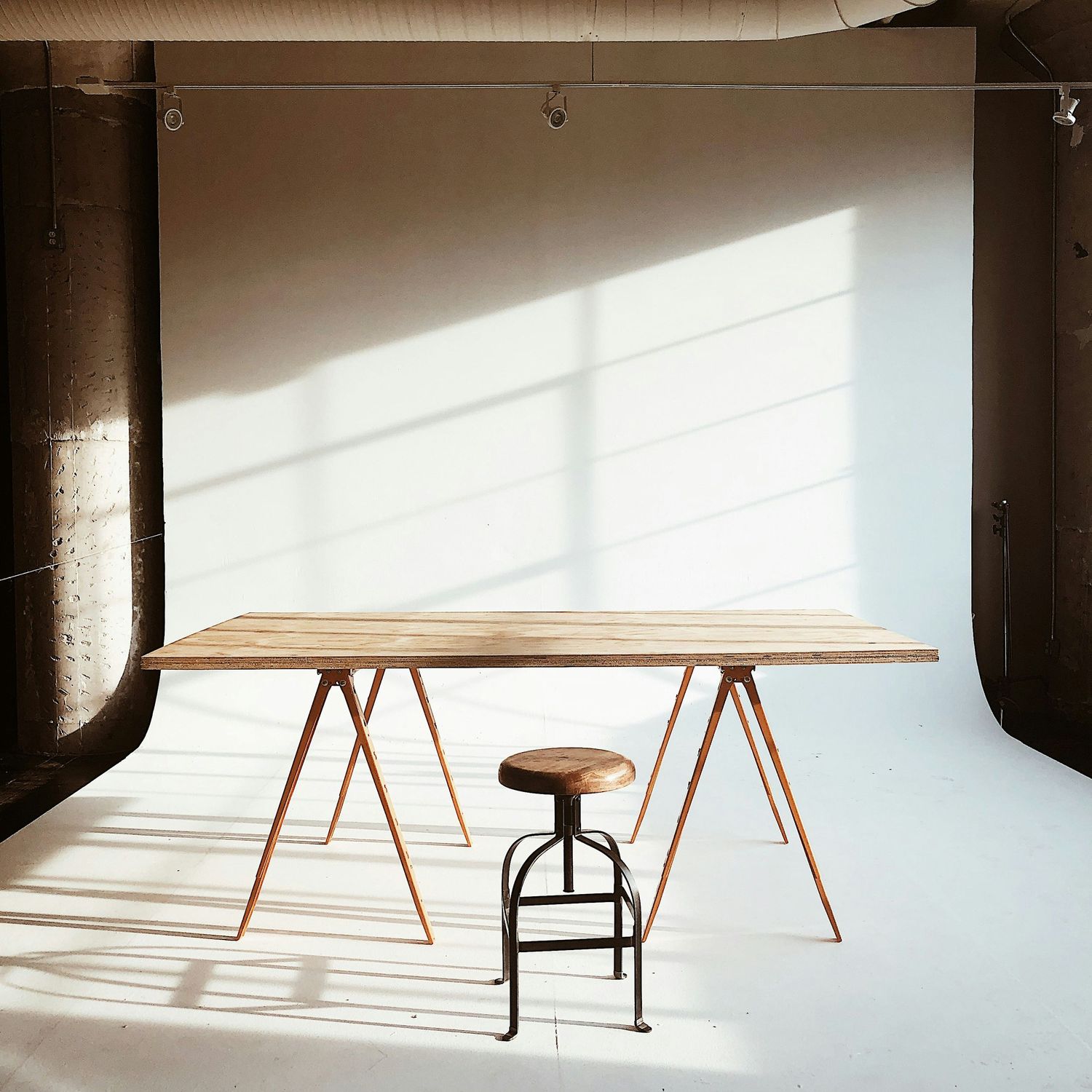 Minimalist photography studio setup with wooden trestle table and vintage industrial stool in natural sunlight.