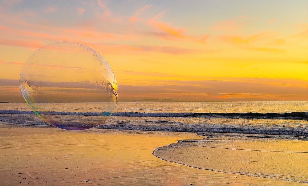 Giant bubble floating above beach waves during a vibrant orange sunset.