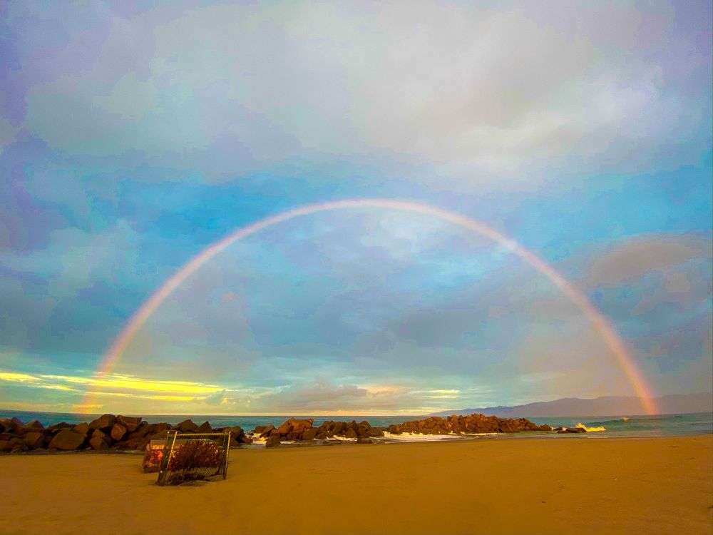 Complete rainbow arches over a sandy beach with buildings and mountains in the distance.
