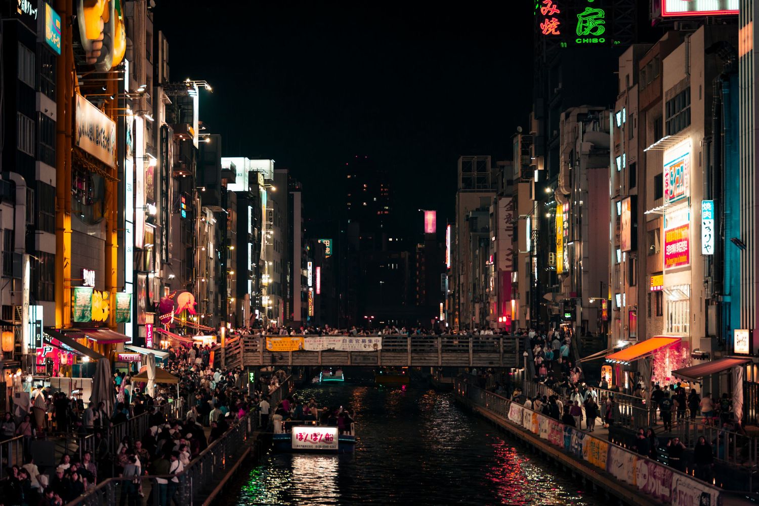 Crowded Dotonbori Canal in Osaka at night with glowing neon signs and storefronts reflecting on the water.