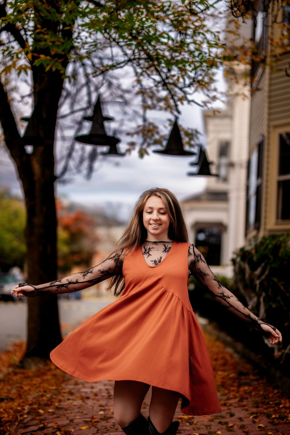 A person in a flowing burnt orange dress twirls playfully among fallen autumn leaves on a tree-lined path.