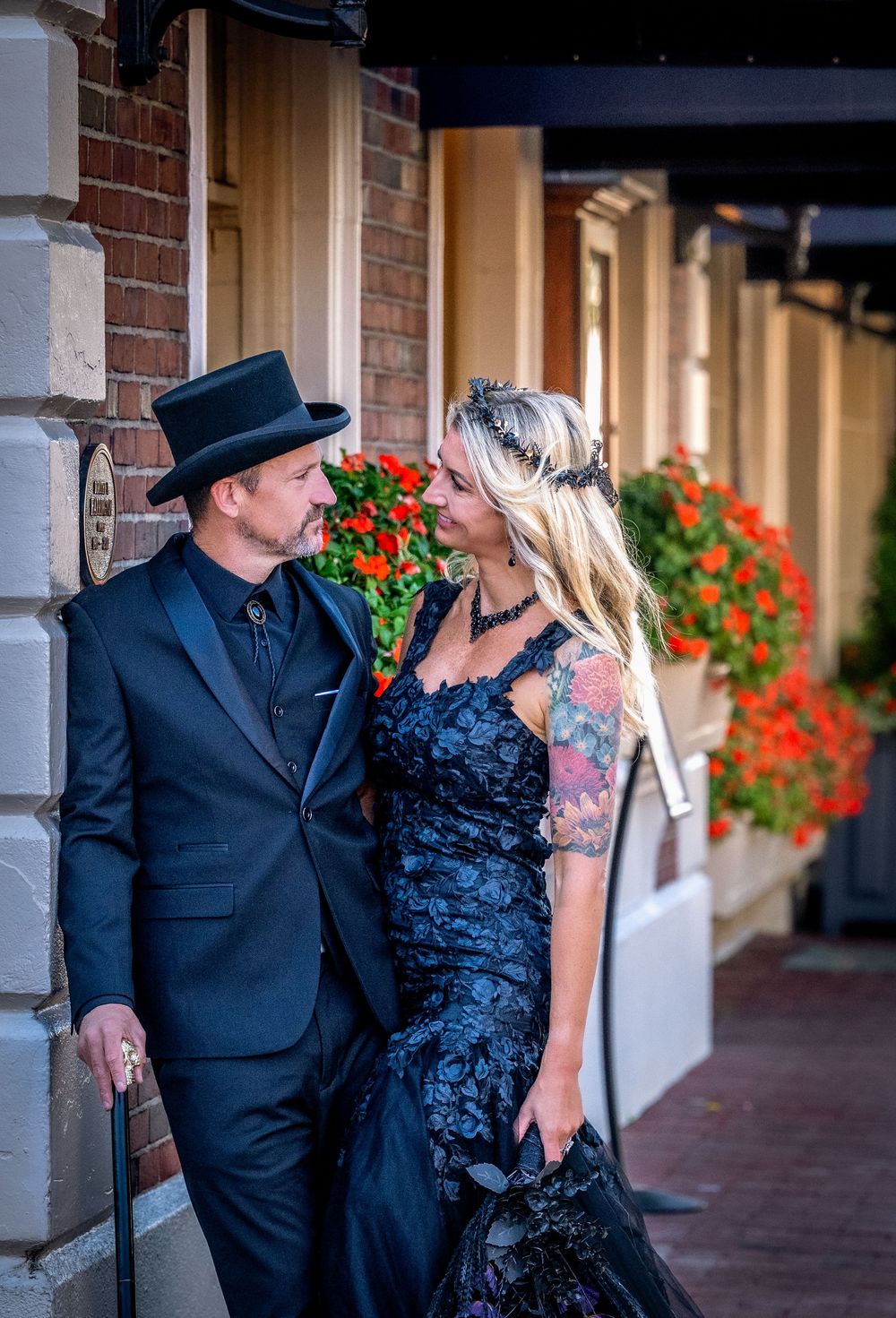 A couple in formal attire shares a romantic moment against a brick wall with red flowers in the background.