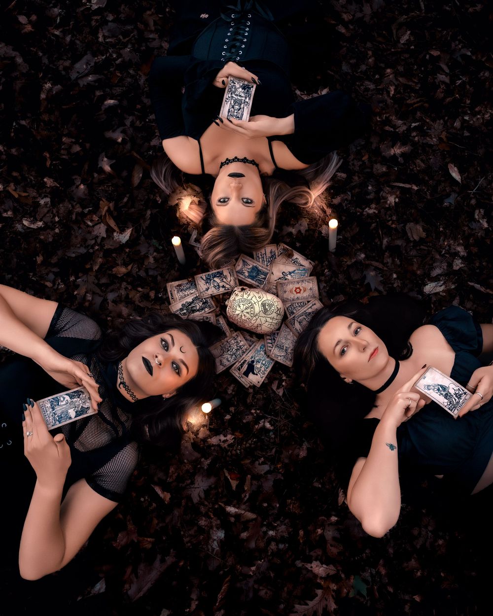 Artistic overhead shots of models lying on dark ground holding playing cards in gothic-inspired fashion.