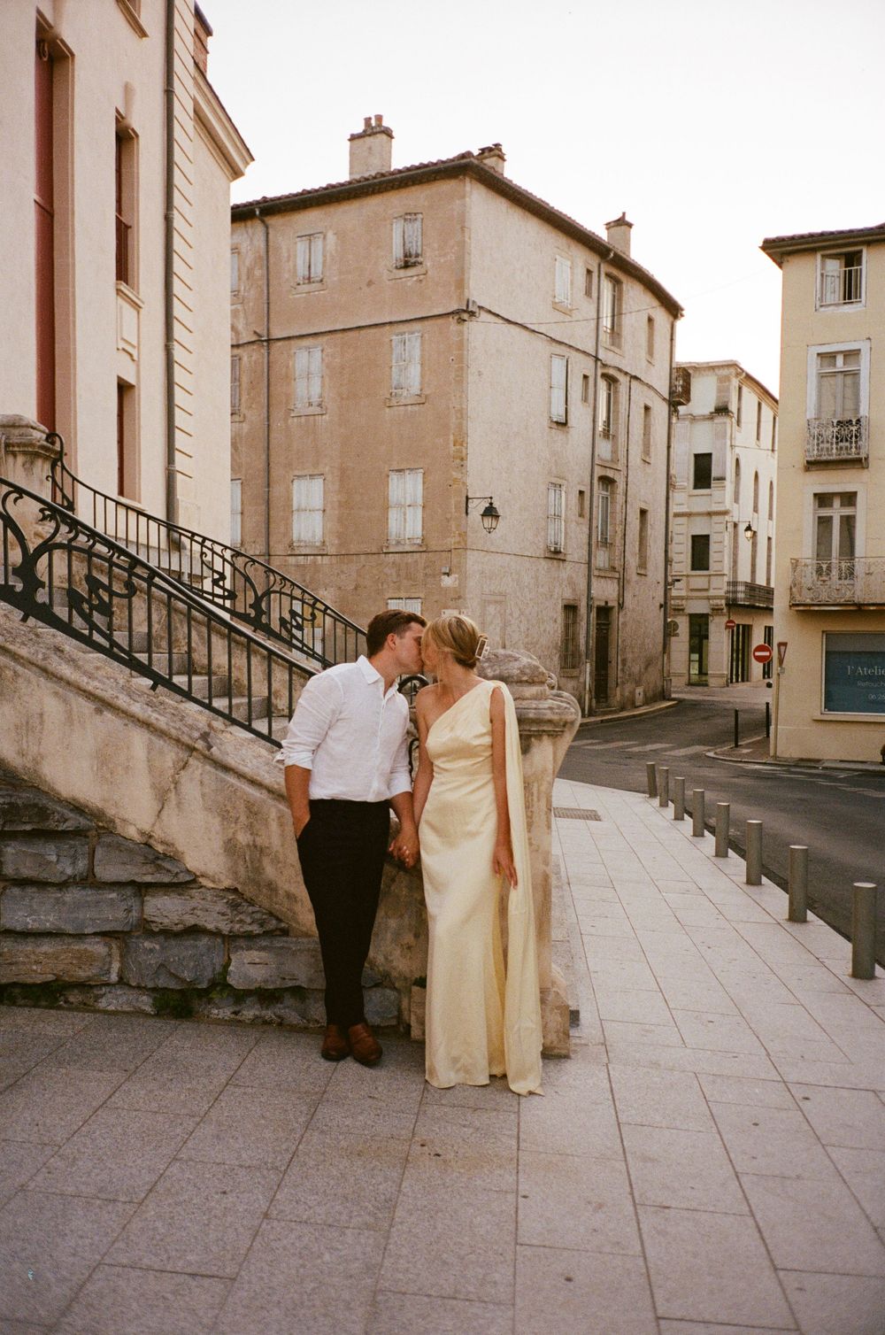 A couple in formal attire sharing a romantic moment on stone steps in front of vintage European buildings.