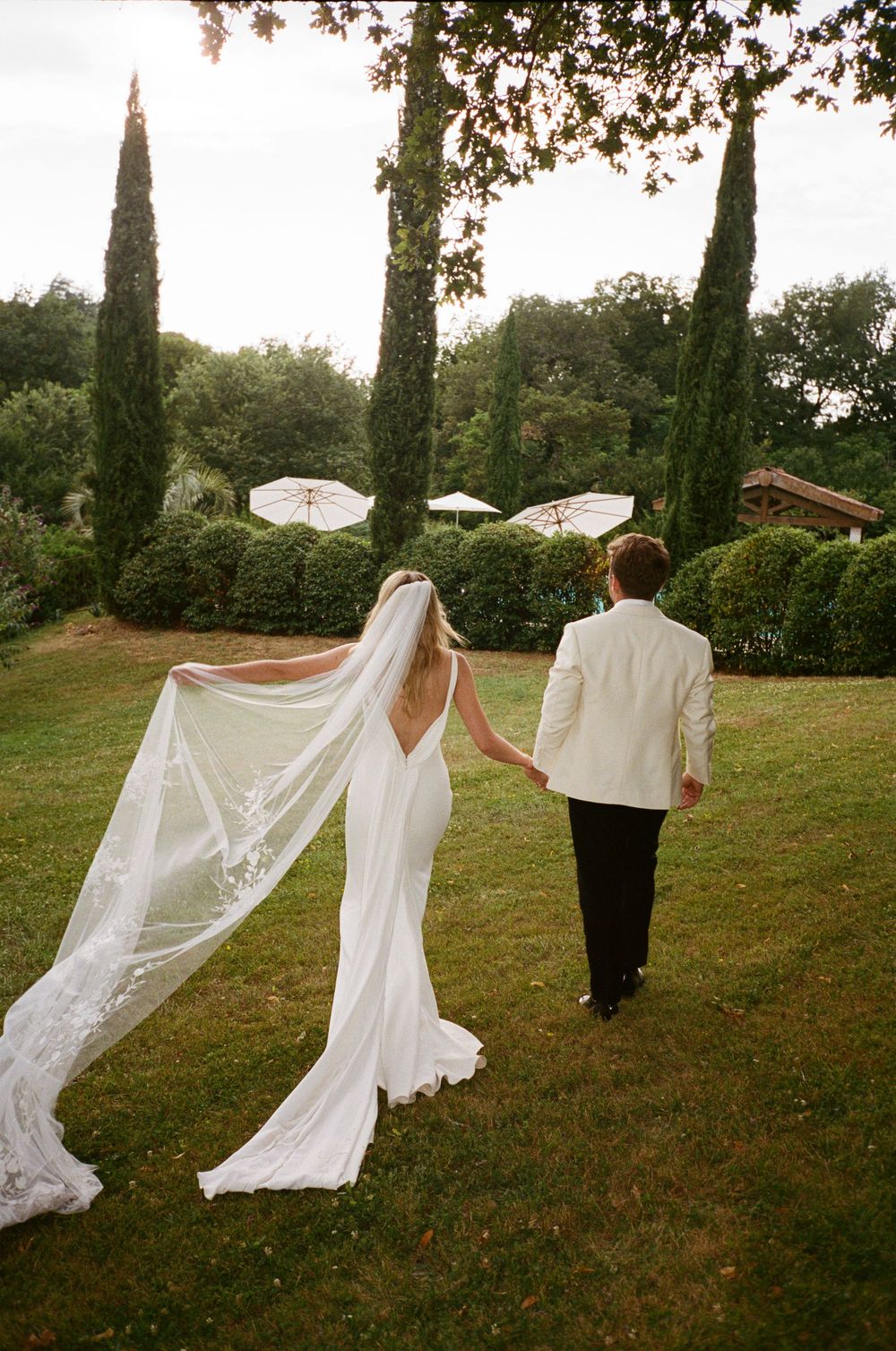 A couple walks on grass with cypress trees and umbrellas in background while a long white veil flows in the breeze.