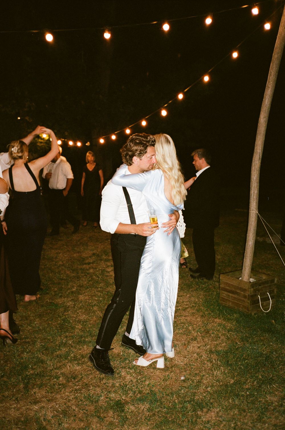 A couple dances together at an outdoor evening reception under warm string lights and a dark night sky.