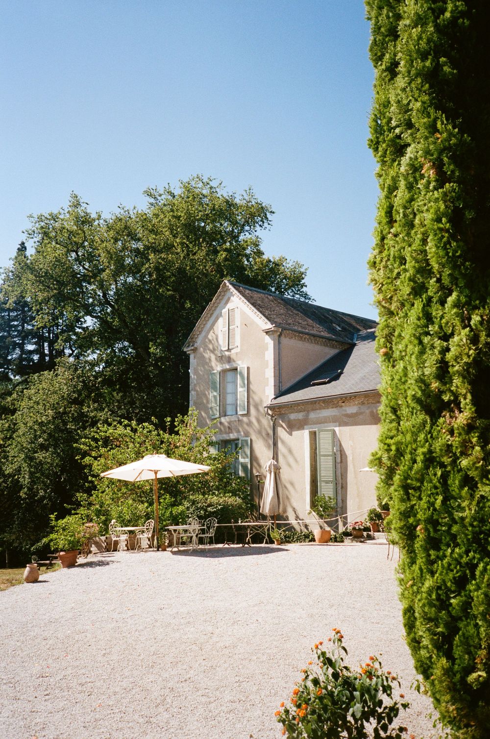 A charming stone villa with gravel driveway features a patio umbrella and cypress trees in the French countryside.