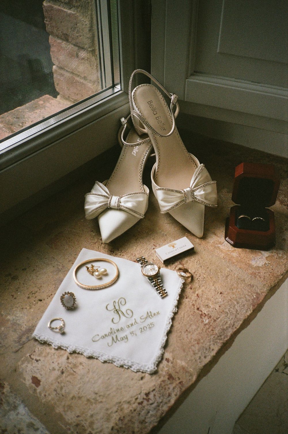 Wedding accessories including white bow heels, rings, and a monogrammed handkerchief arranged on a windowsill.
