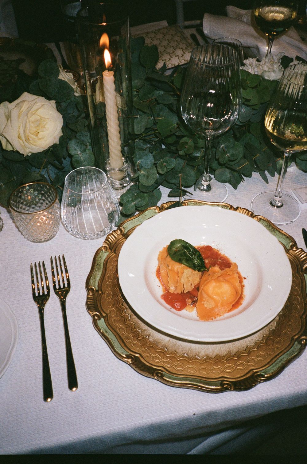 An elegant place setting with candle, wine glasses and gourmet meal on a gold-rimmed plate.