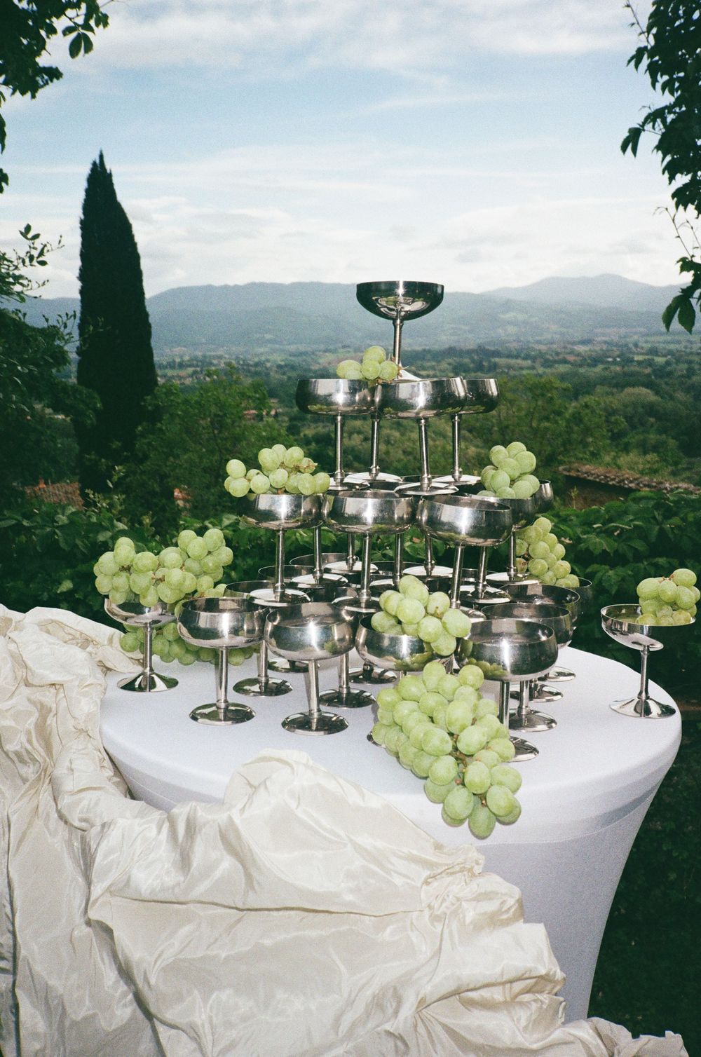Elegant buffet table with silver champagne stands and fresh green grapes overlooking a Tuscan landscape.Tuscany Wedding Photographer