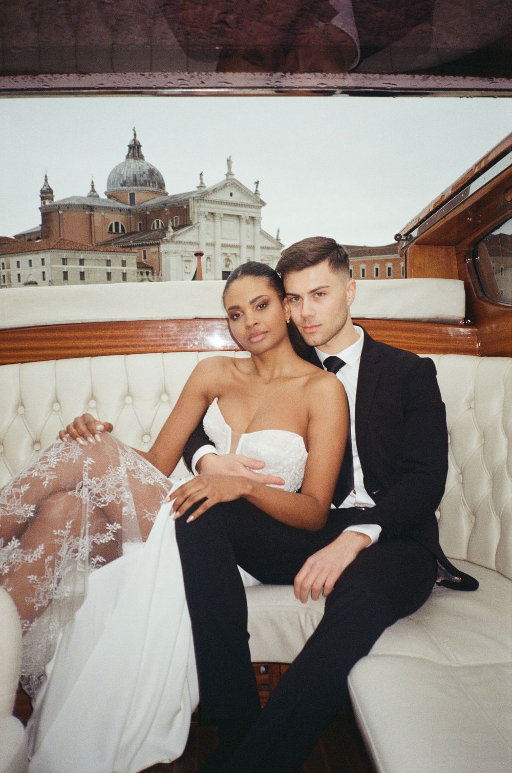 An elegant couple in wedding attire pose together on a luxurious white settee with ornate architecture in the background.