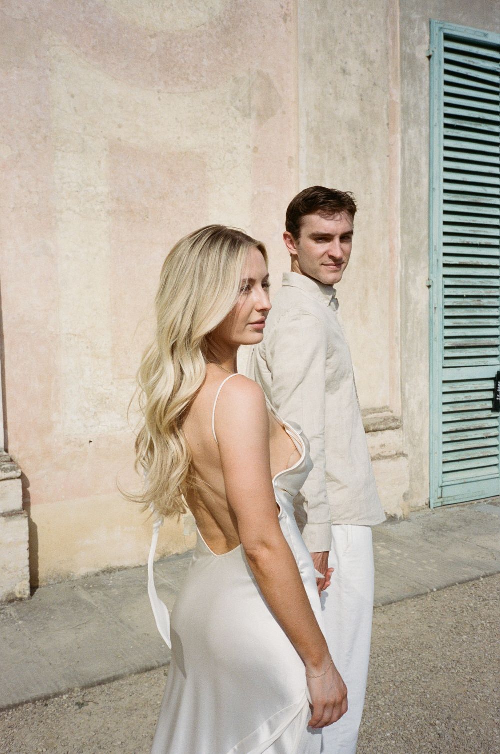A couple in elegant white attire poses against a rustic wall with blue shutters in a romantic outdoor setting.