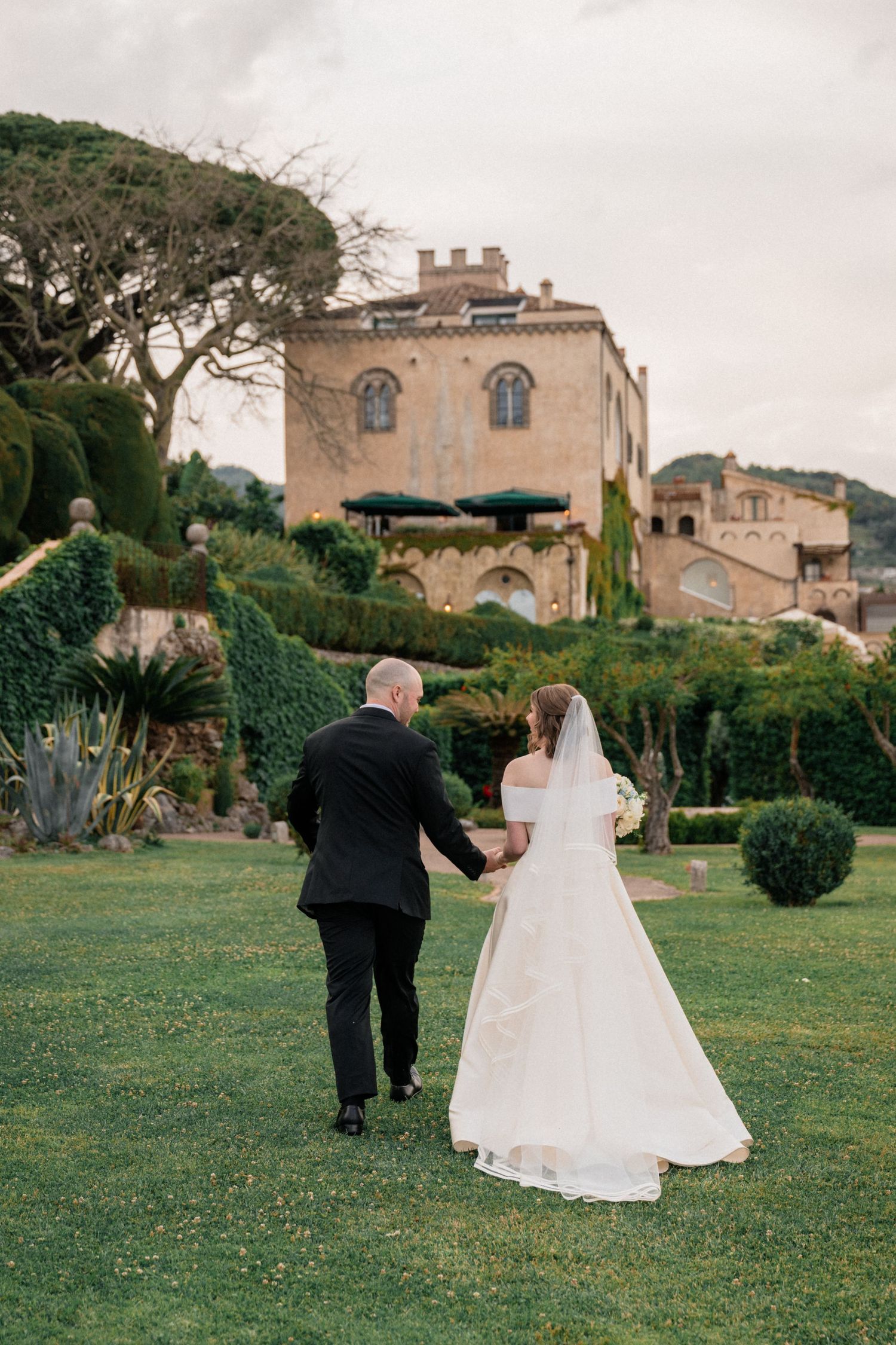 A couple walks together on a manicured lawn with an elegant Italian villa and gardens in the background.