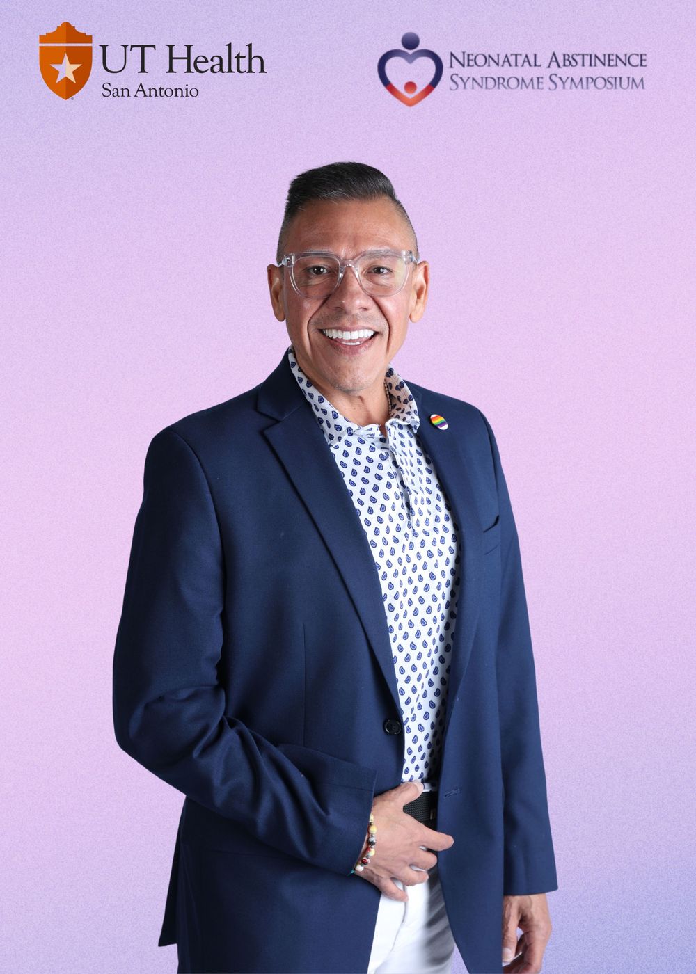 Professional wearing a navy suit and polka dot shirt stands against light purple background at healthcare event.