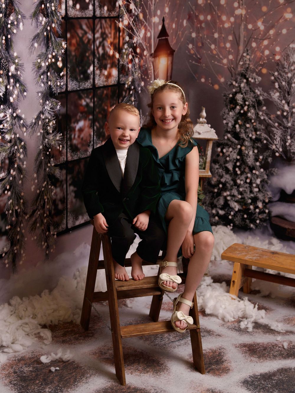 Children sitting on wooden benches in a winter wonderland photo studio setting with snowy trees and bokeh lights.