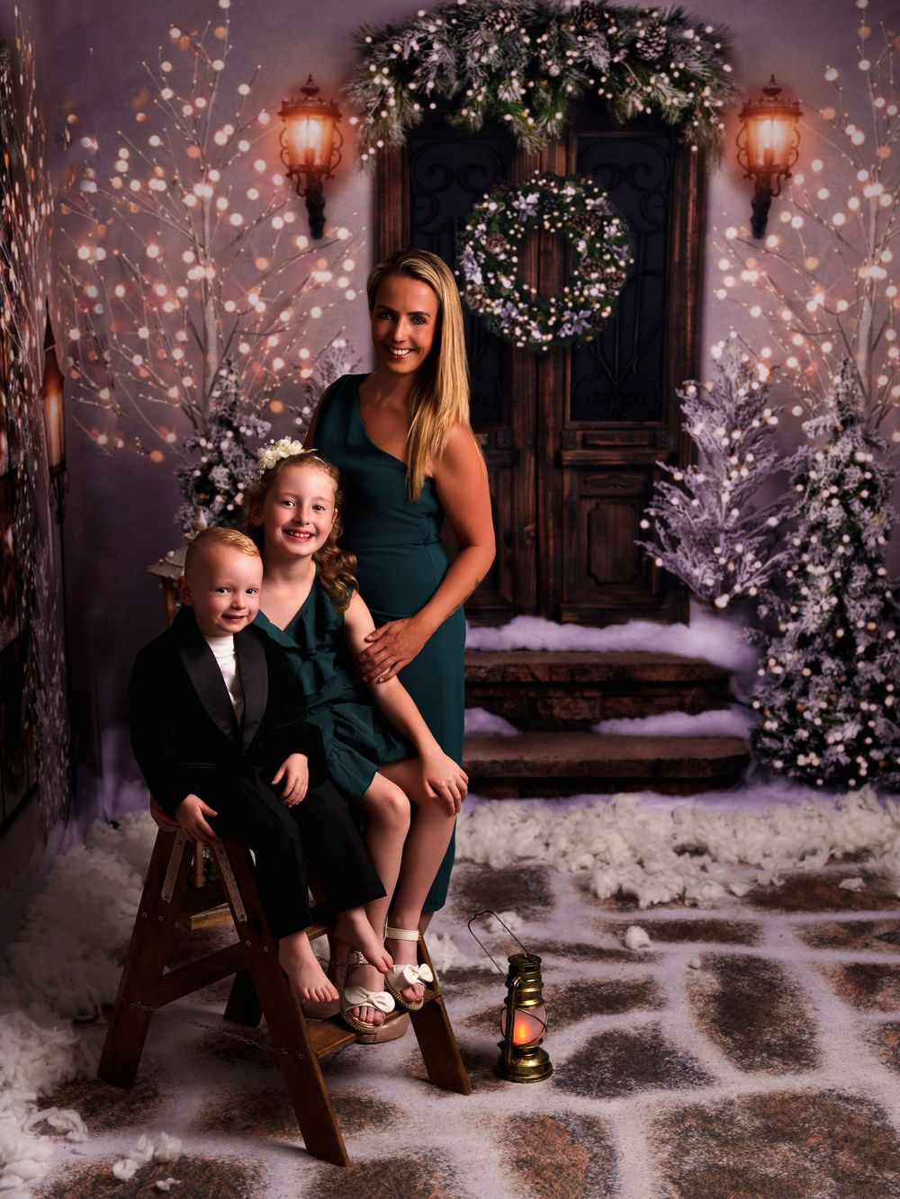 Festive winter holiday portrait setup with glowing lanterns, decorated door, wreath and snow-covered steps in magical lighting.