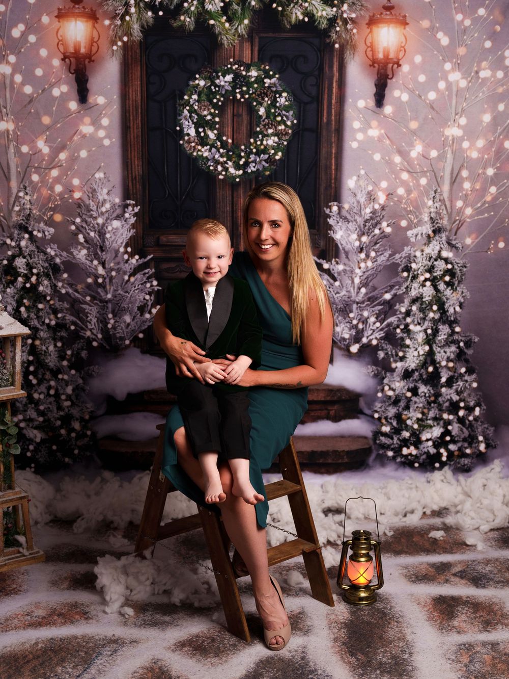A cozy winter portrait setup with glowing lanterns, snow-covered trees, and a rustic wooden chair against a dark backdrop.