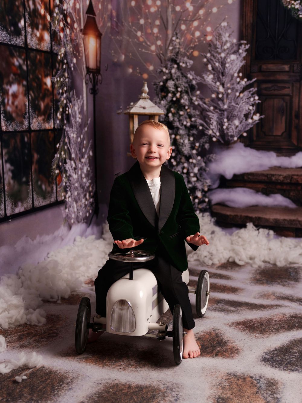 A child in formal attire sits in a winter wonderland setting with snow and twinkling lights in a photo studio.