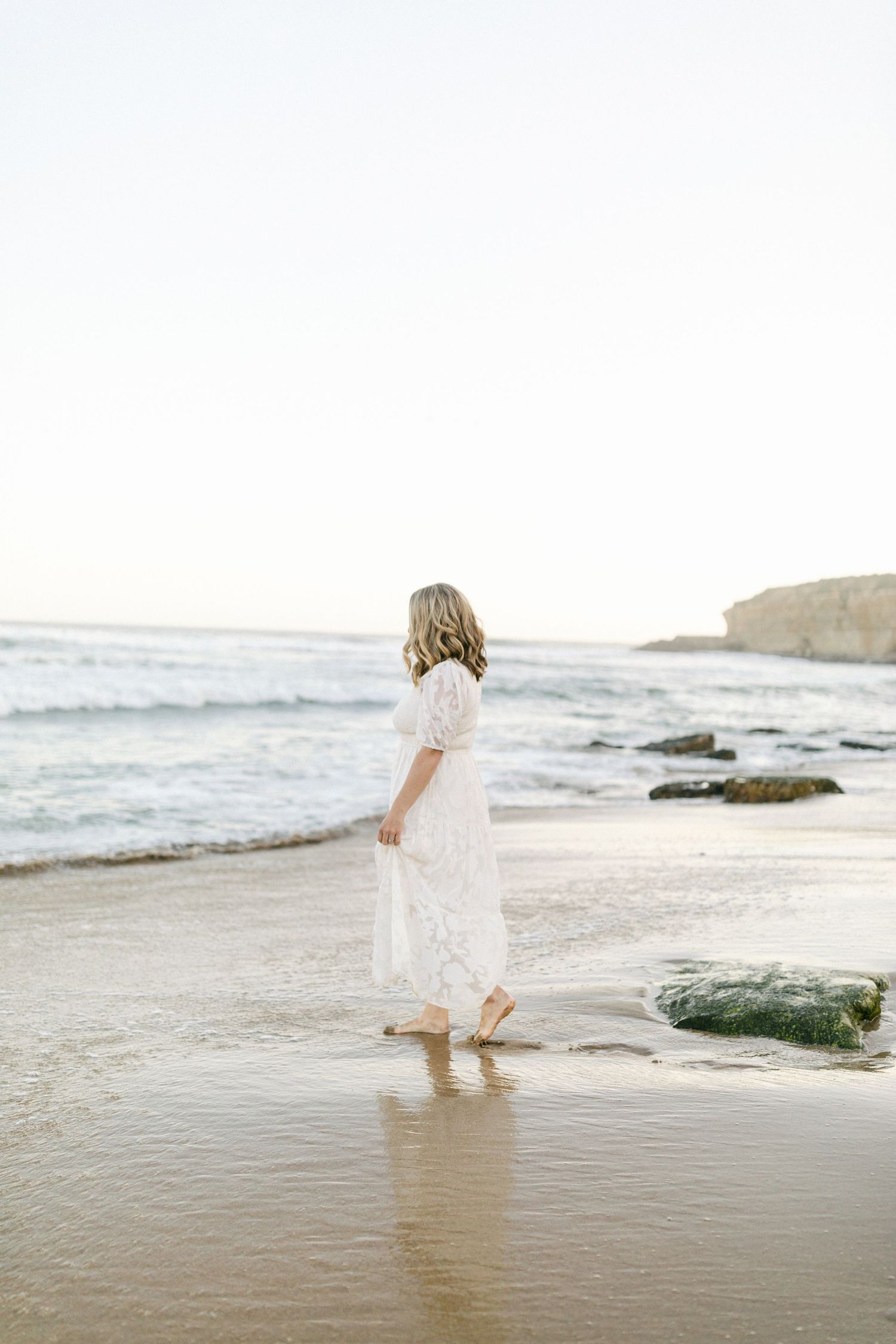 A person in a flowing white dress stands at the edge of the ocean waves on a sunny beach captured by Chelsee Rawe Photography.