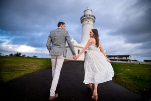 Couple in wedding attire walk along dramatic hilltop path with lighthouse and stormy sky in background on their proposal / engagement day.