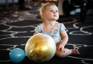 Baby in light colored outfit lying on patterned floor with colorful balloons.